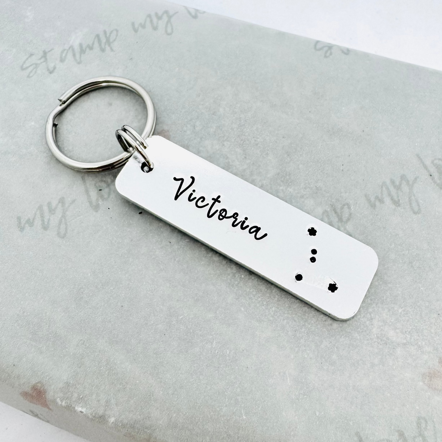 Hand stamped aluminium keyring personalised with the name Victoria and a zodiac constellation design, attached to a stainless steel keyring and displayed on eco-friendly Stamp My Love branded packaging.