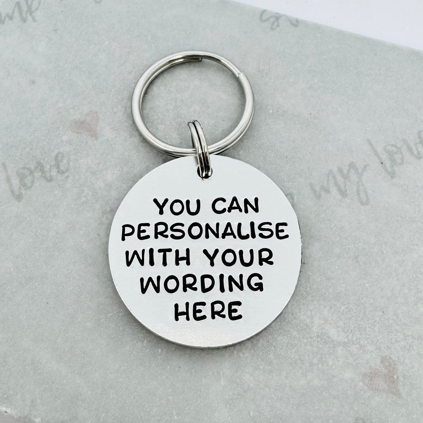 Round personalised hand stamped aluminium keyring with custom wording example on a white background with silver keyring attachment.