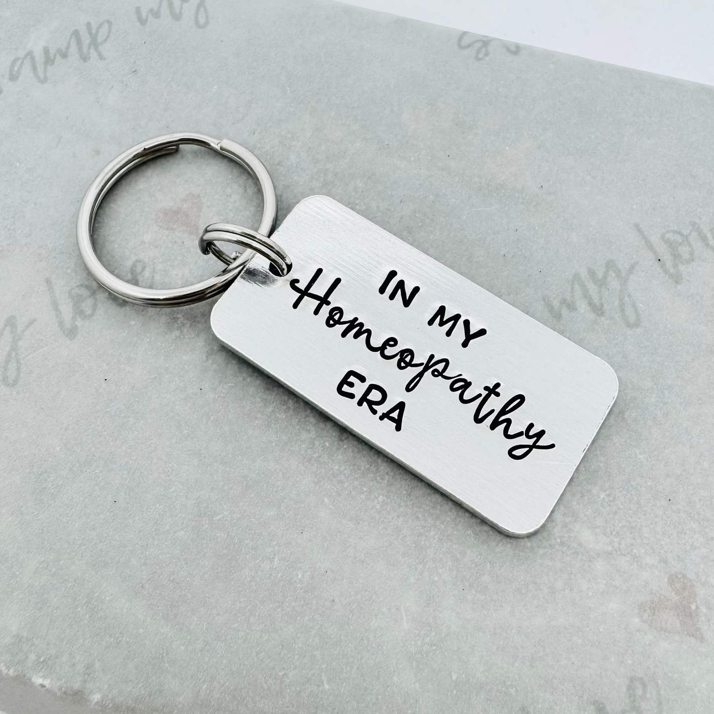 Hand stamped aluminium keyring with the phrase “In My Homeopathy Era” in black lettering, displayed on eco-friendly branded packaging with stainless steel keyring attachment.