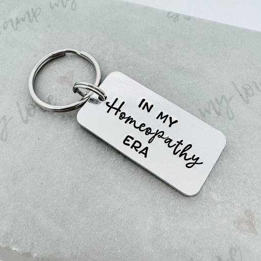 Hand stamped aluminium keyring with the phrase “In My Homeopathy Era” in black lettering, displayed on eco-friendly branded packaging with stainless steel keyring attachment.