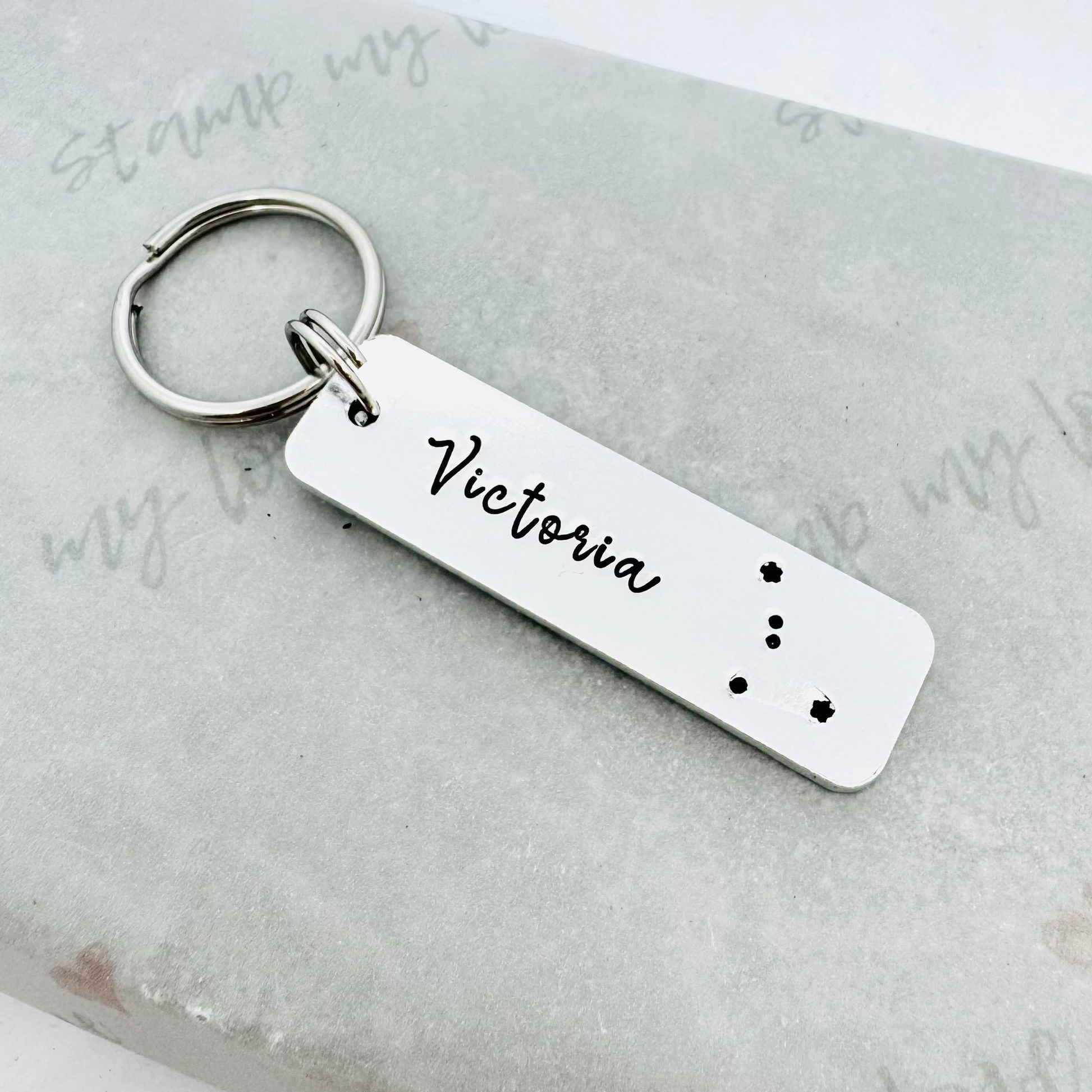 Hand stamped aluminium keyring personalised with the name Victoria and a zodiac constellation design, attached to a stainless steel keyring and displayed on eco-friendly Stamp My Love branded packaging.