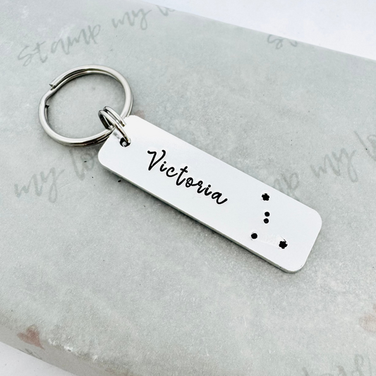 Hand stamped aluminium keyring personalised with the name Victoria and a zodiac constellation design, attached to a stainless steel keyring and displayed on eco-friendly Stamp My Love branded packaging.